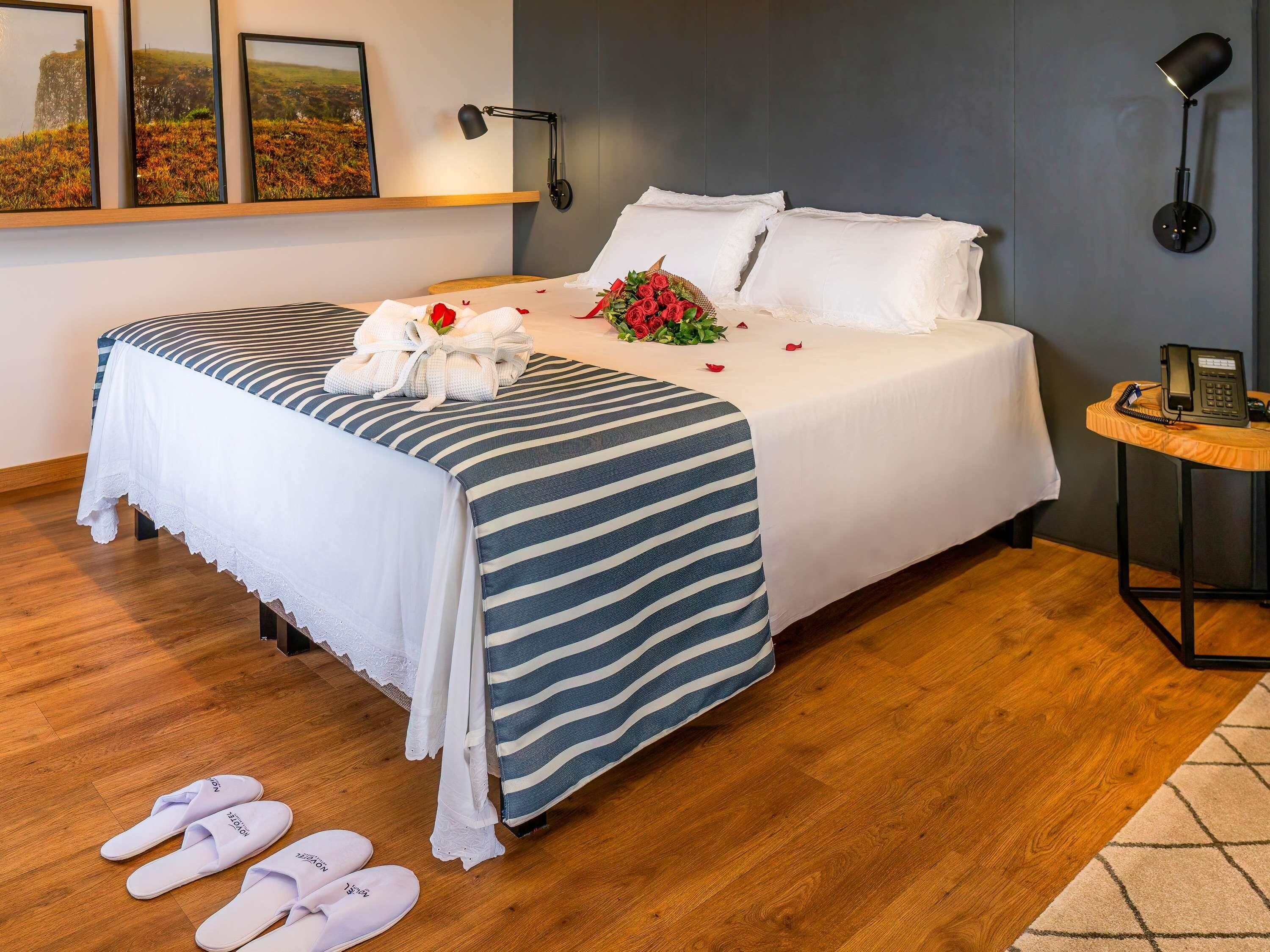 Una cama bien hecha con una colcha a rayas, un ramo de flores y un par de zapatillas en el suelo.
