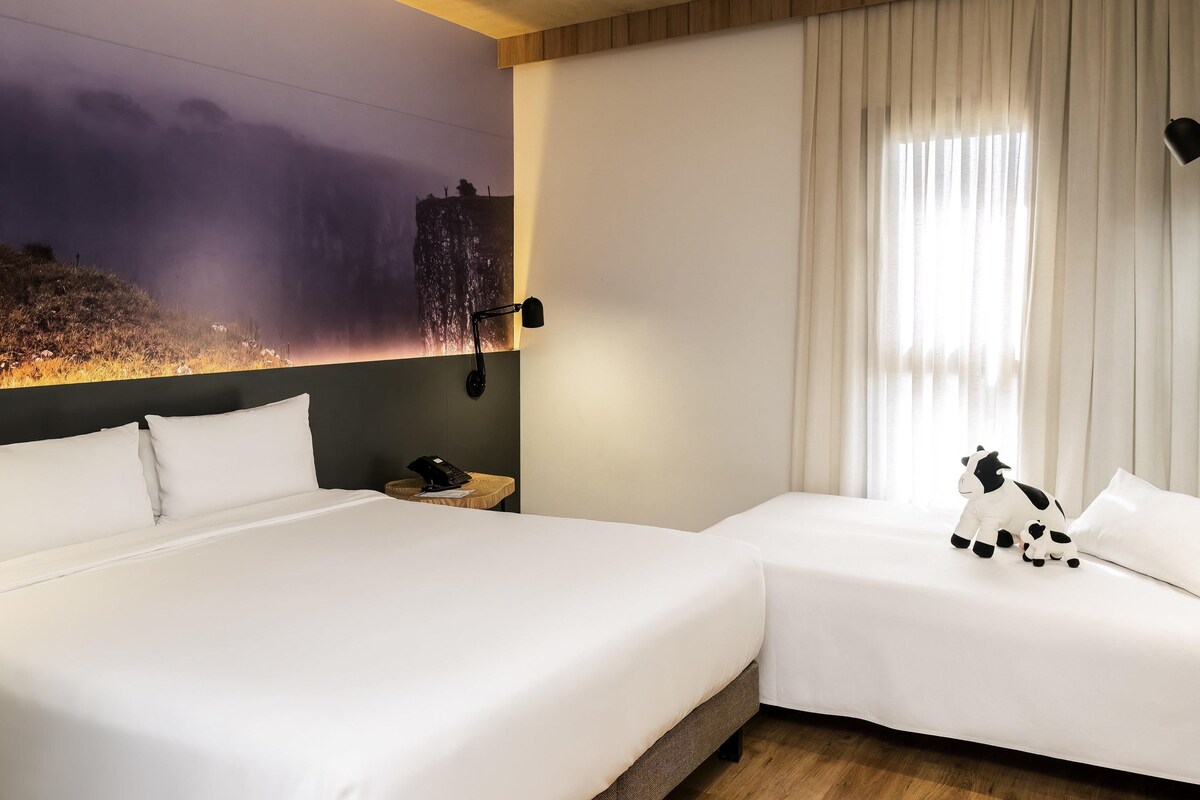 Habitación de hotel con dos camas, una mesita de noche, una lámpara, un mural en la pared y una ventana con cortinas.