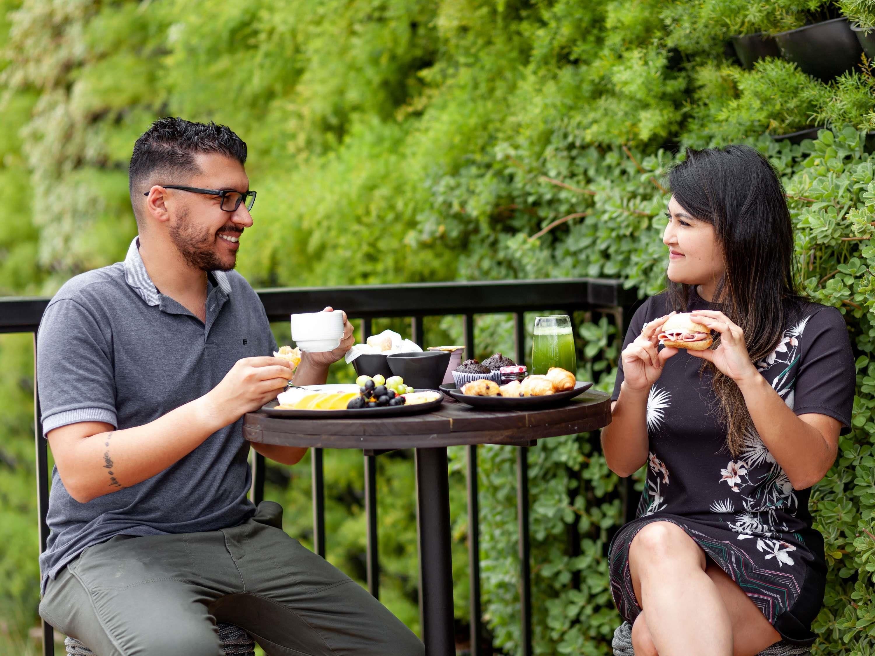 Two people sitting at an outdoor table with food and drinks, surrounded by greenery.