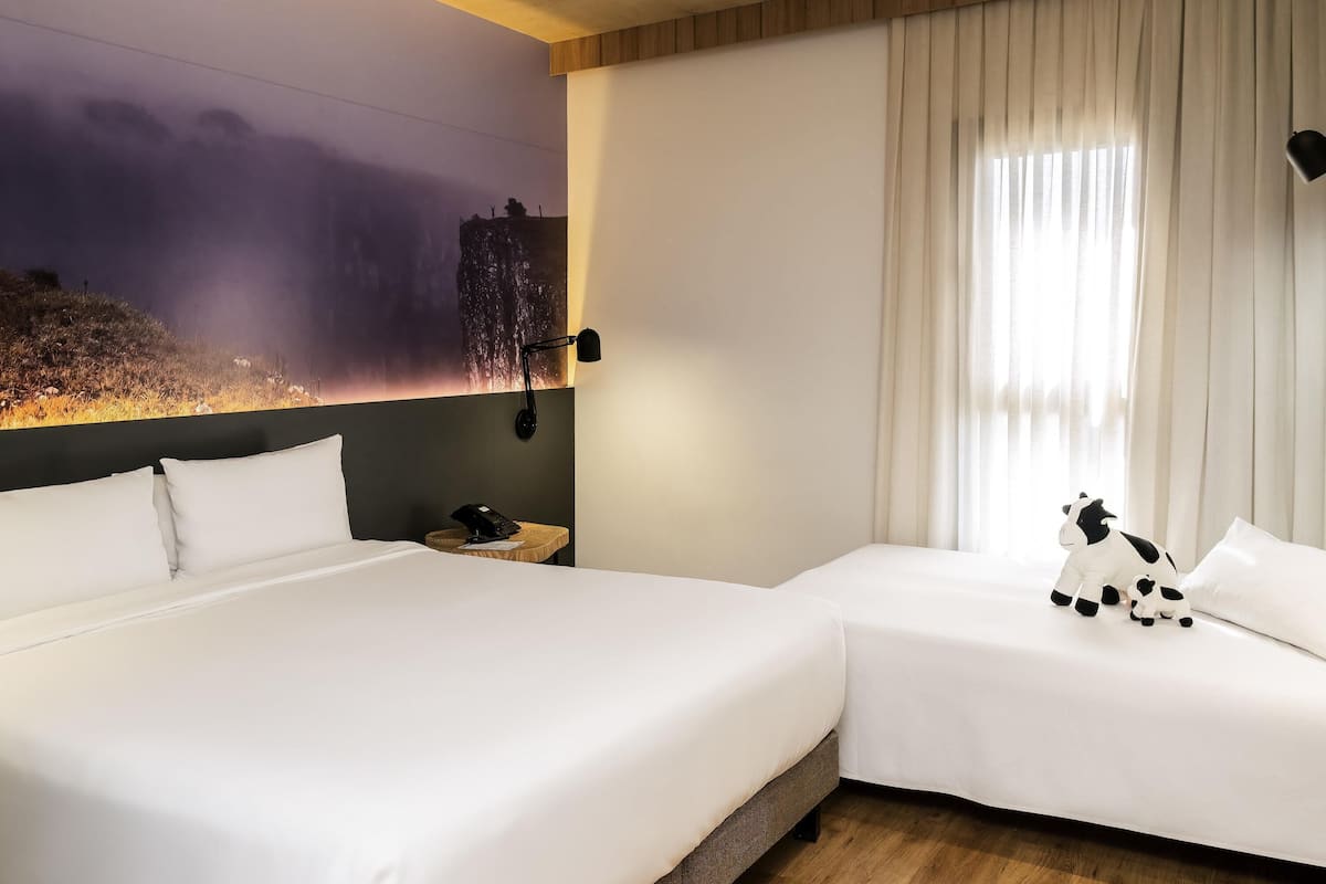Hotellihuone, jossa on kaksi sänkyä, yöpöytä, lamppu, seinämaalaus ja ikkuna, jossa on verhot.
