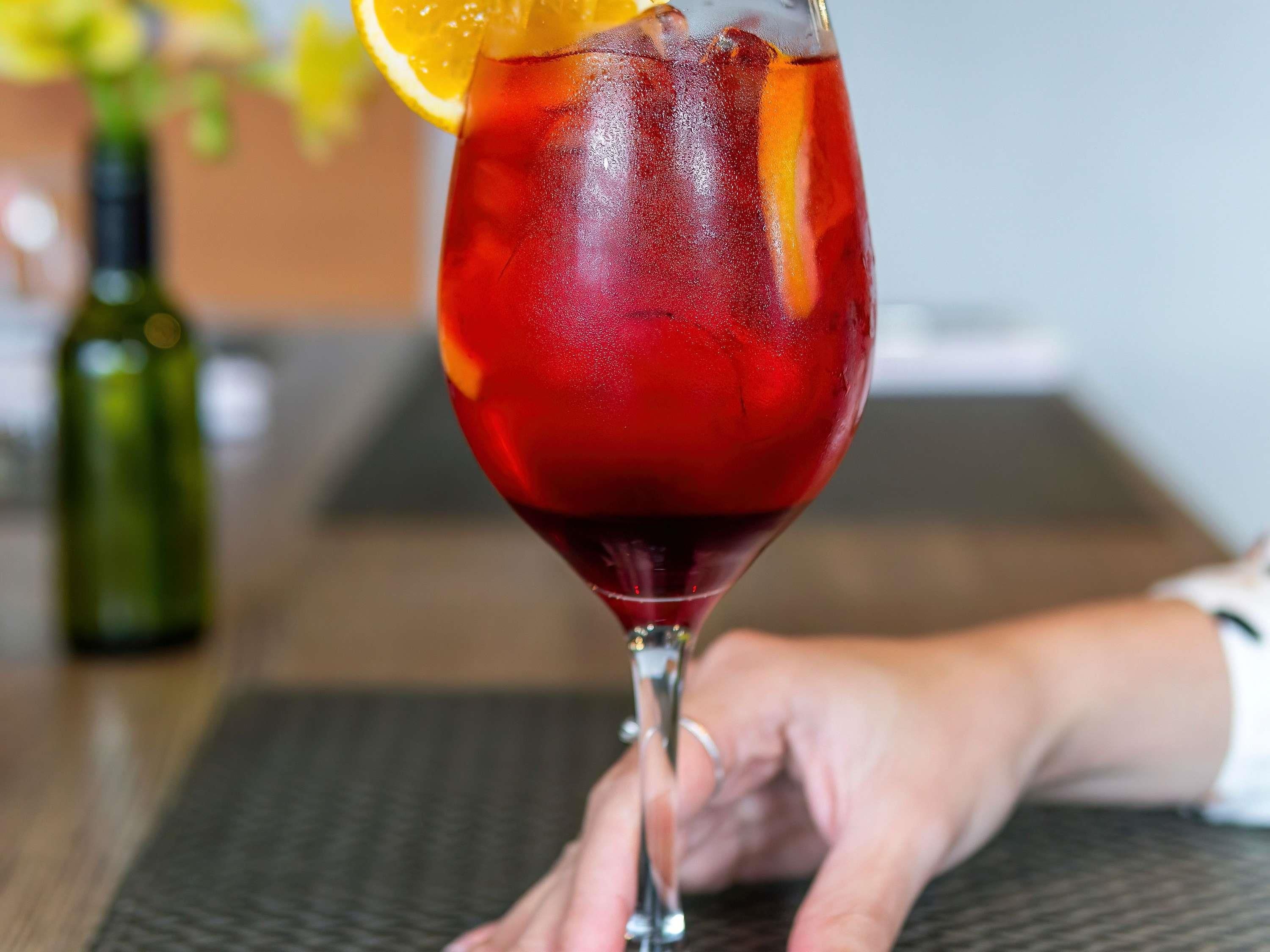 Una mano sostiene un vaso con una bebida roja y una rodaja de naranja.