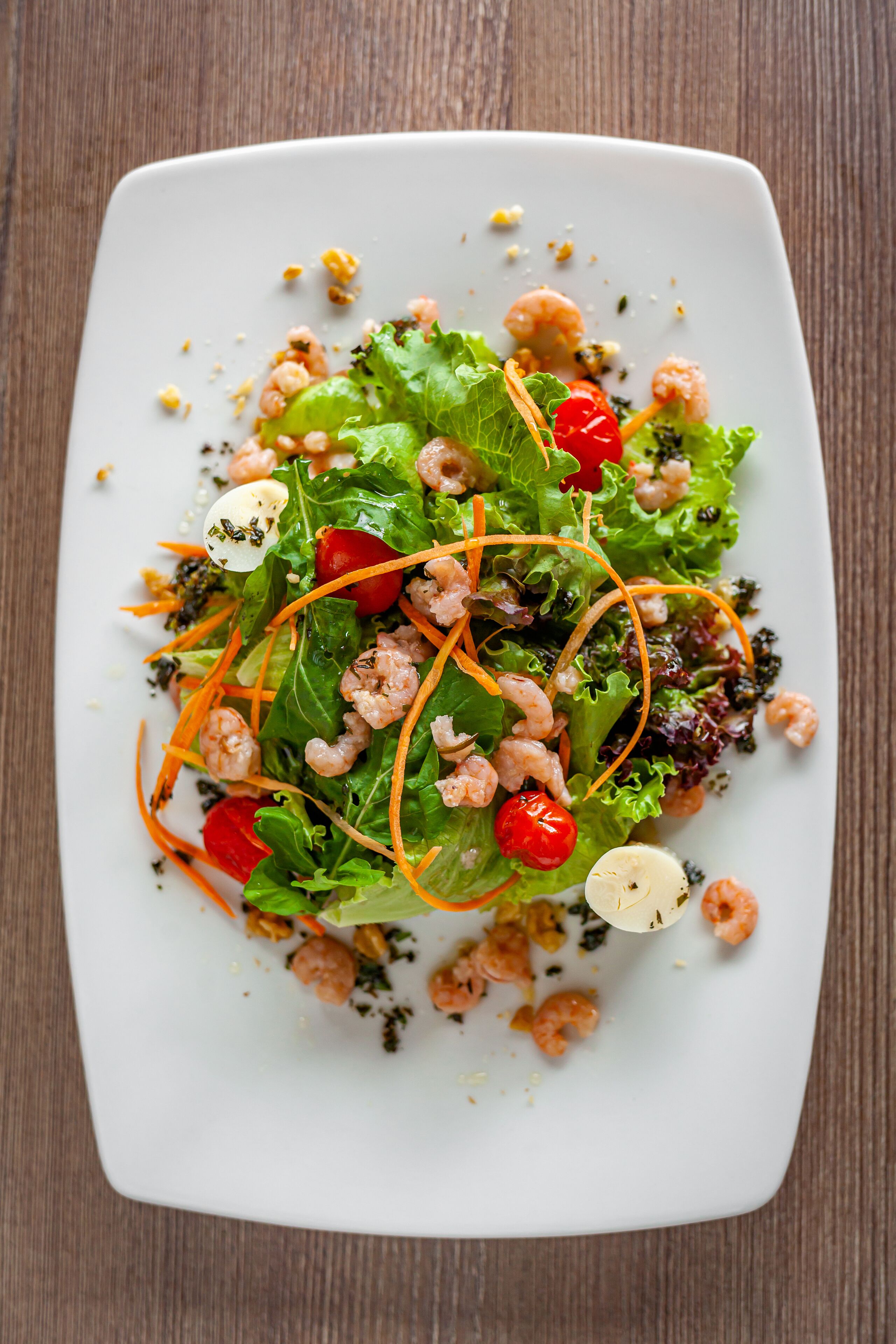 Un plato rectangular blanco con una ensalada de lechuga, tomates cherry, zanahorias y camarones.