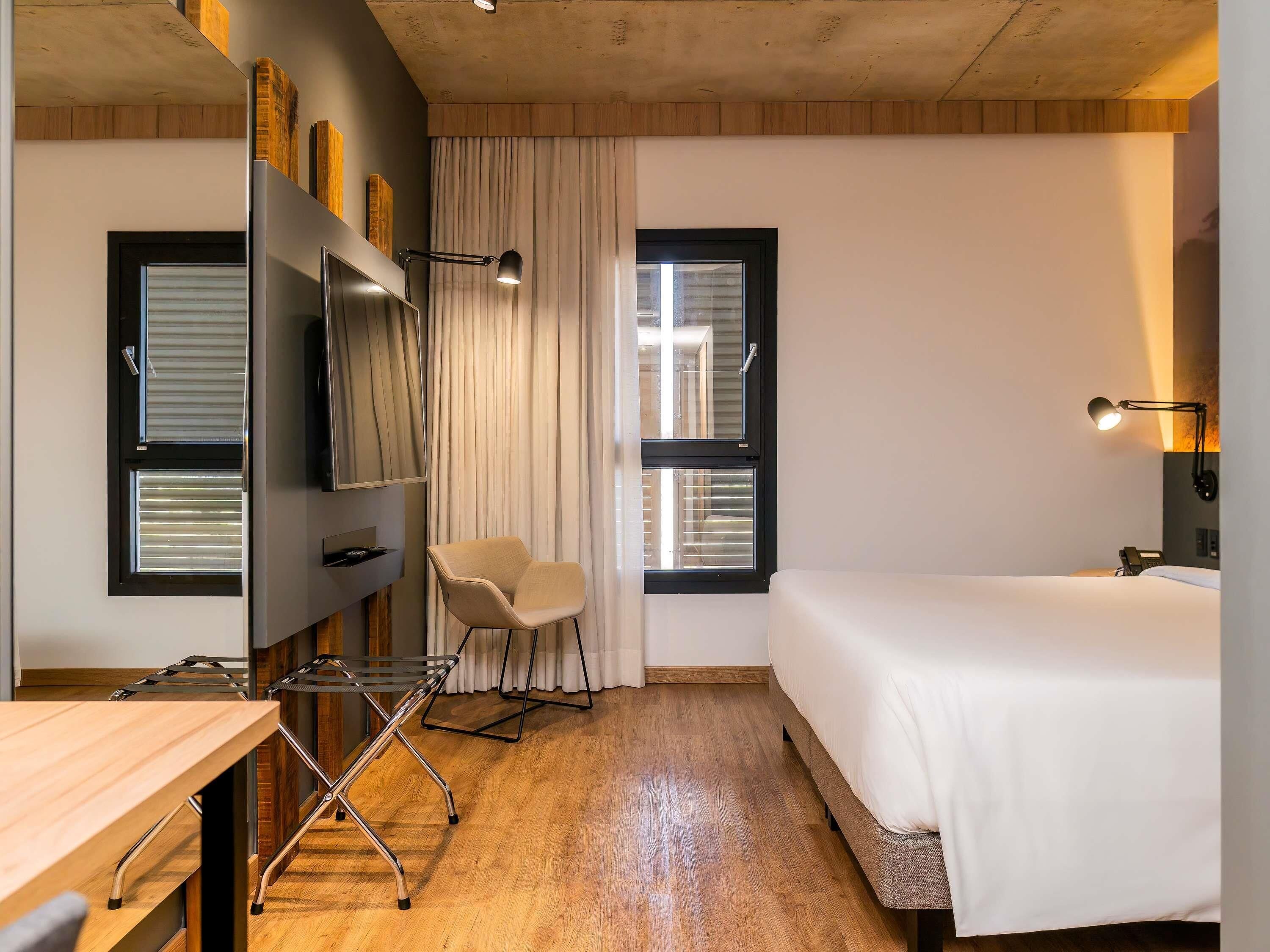 A modern hotel room with a bed, a desk, a chair, a TV, and a window with blinds.