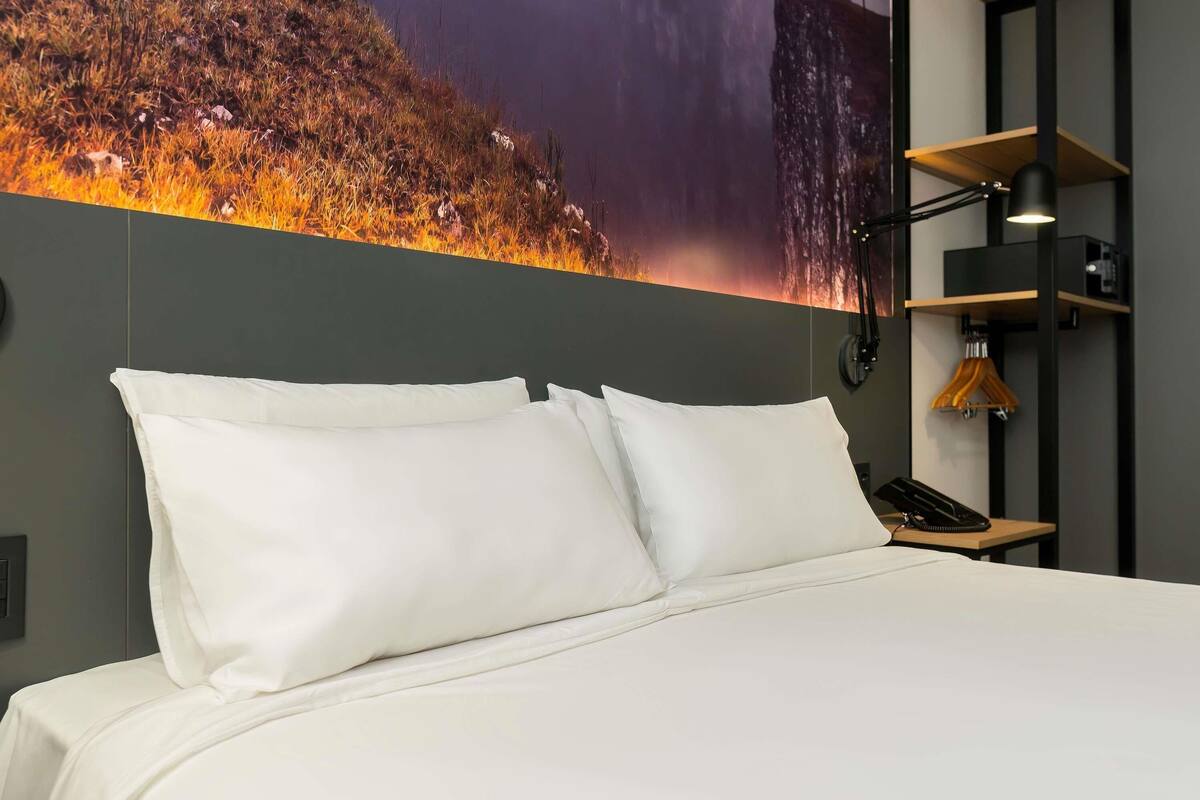 A hotel room with a large bed, white pillows, a wall-mounted phone, a lamp, and a wooden shelf with a hanger.