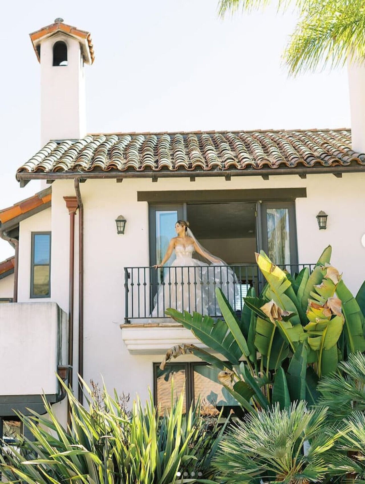 A two-story house with a balcony, a small tower-like structure on the roof, and lush tropical plants in the foreground.