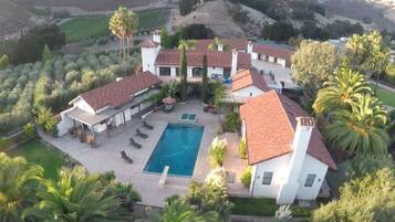 A large house with a swimming pool, surrounded by palm trees and a hilly landscape.