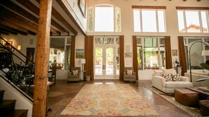 A spacious living room with a large rug, wooden beams, and a view through a glass door to the outside.