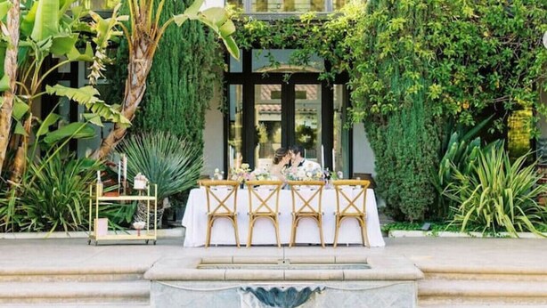 A poolside wedding setup with a table and chairs, surrounded by lush greenery and a tiled-roof building.