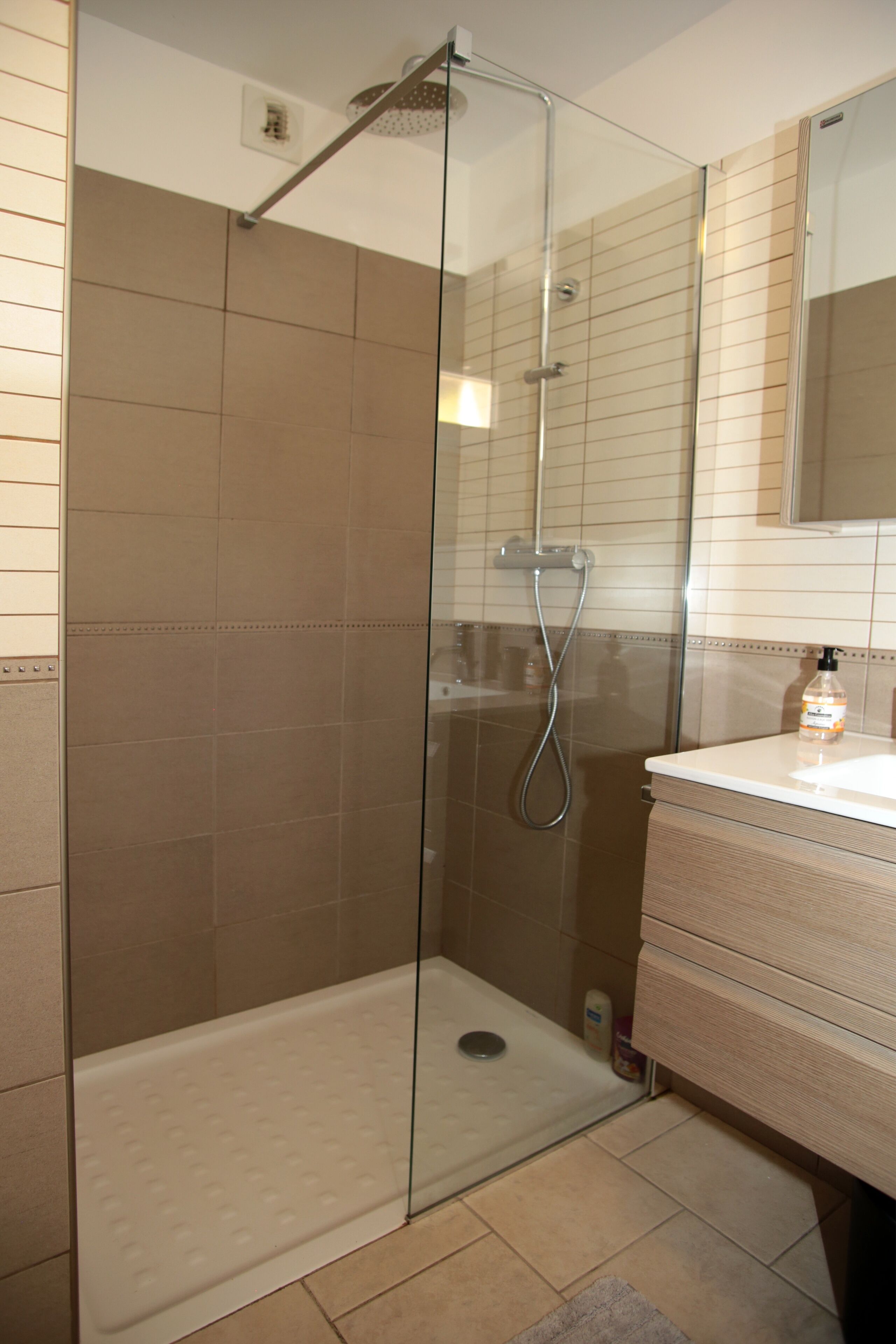 A glass-enclosed shower with a white floor, a sink with a mirror above it, and beige tiles on the walls.