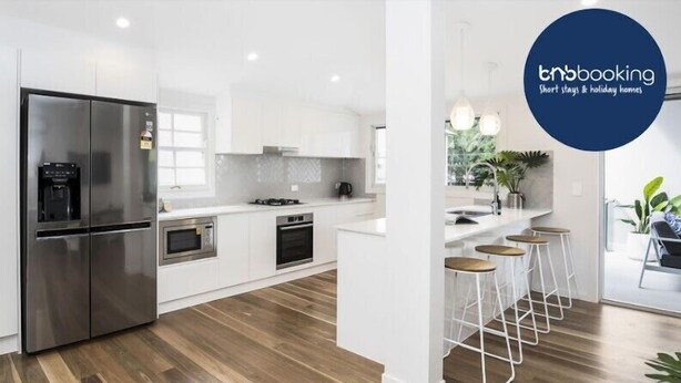 A modern kitchen with white cabinetry, stainless steel appliances, and a wooden floor. There are bar stools near a window with a view of greenery.