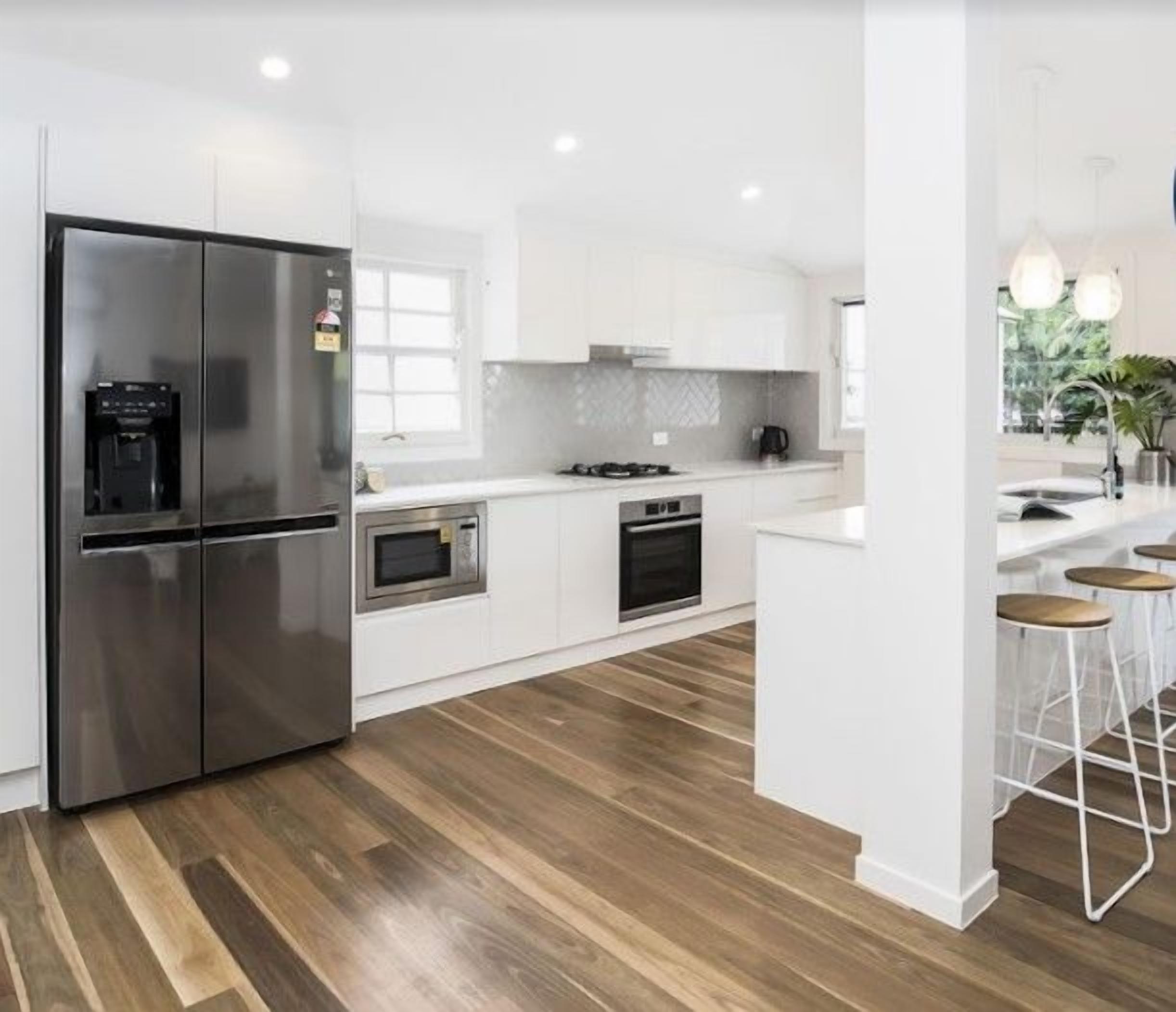 A modern kitchen with white cabinetry, stainless steel appliances, and a wooden floor. There are bar stools near a window with a view of greenery.