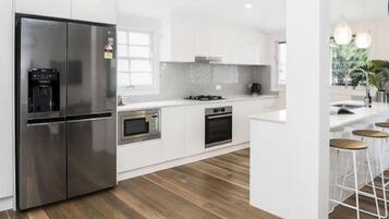 A modern kitchen with white cabinetry, stainless steel appliances, and a wooden floor. There are bar stools near a window with a view of greenery.
