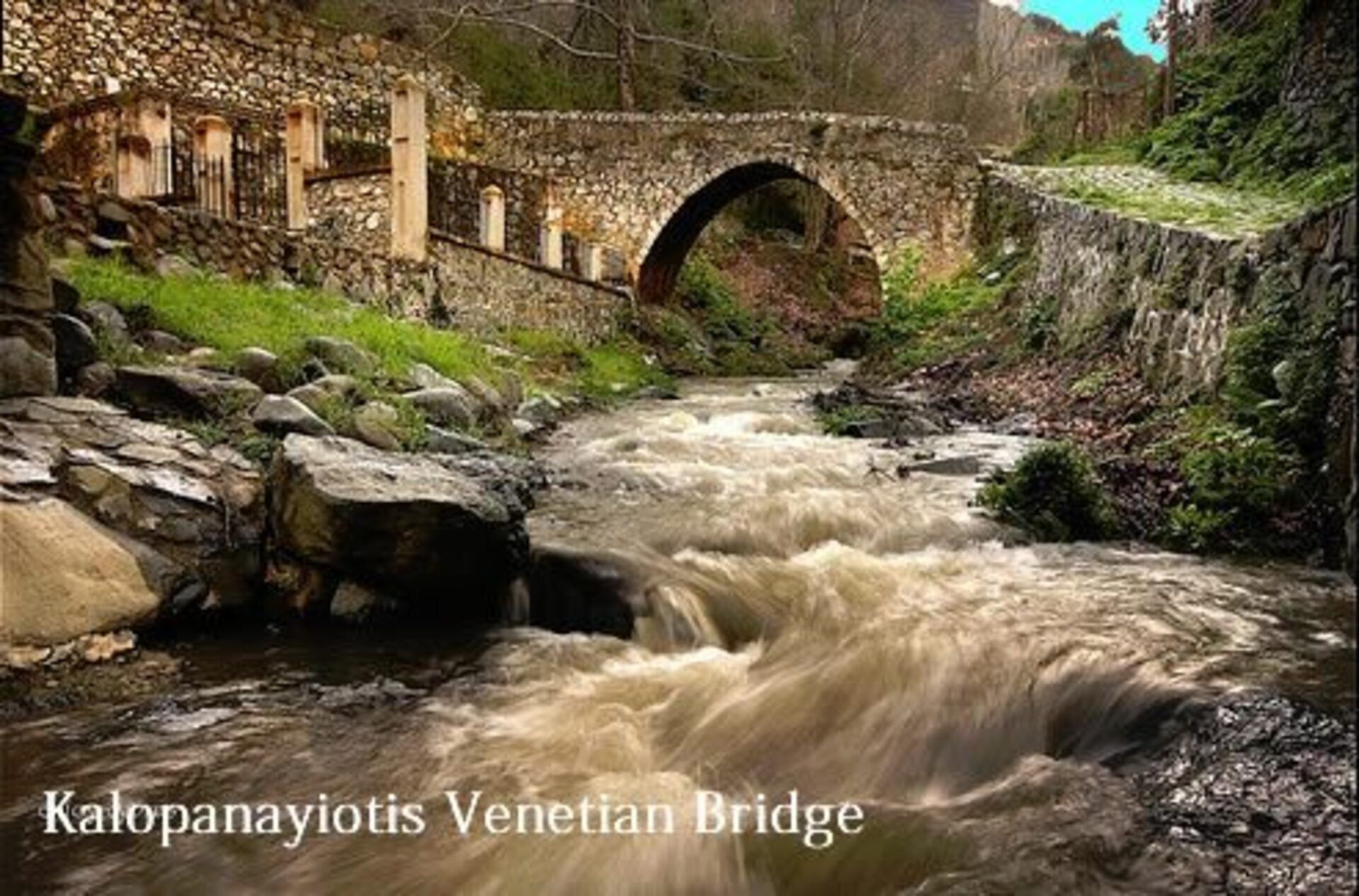 A stone bridge over a flowing river with a stone wall on one side and a grassy area on the other.