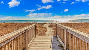 Ein hölzerner Steg führt zu einem Strand, im Hintergrund ein klarer Himmel und das Meer.