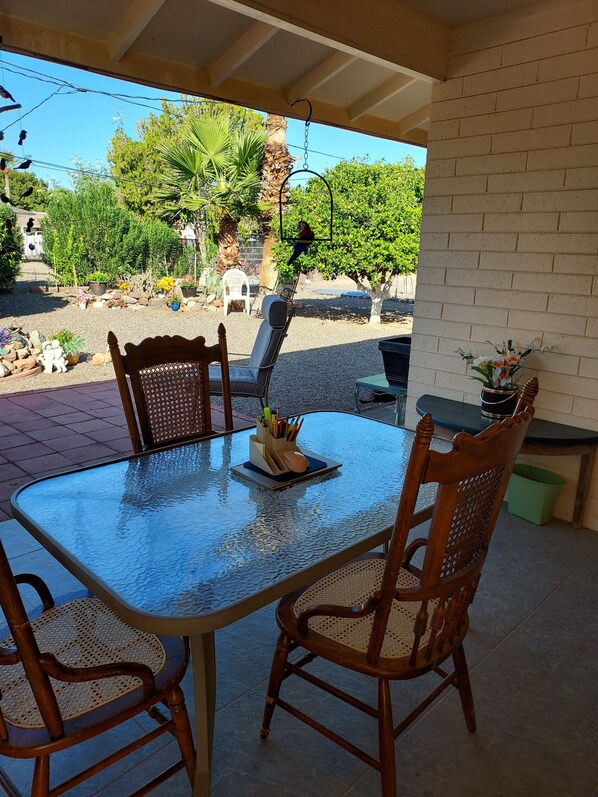 A glass-top table with two wooden chairs, a small potted plant, and a metal lamp on a patio with a brick wall and greenery in the background.