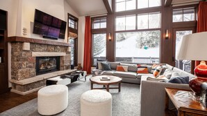 A cozy living room with a stone fireplace, a flat-screen TV mounted above it, a large window with a view of a snowy landscape, a comfortable sofa, and a coffee table. - Luxurious Ski-in Ski-out Home in Arrowhead community (Edwards)