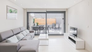 Un salon moderne avec un canapé gris, un téléviseur à écran plat et une porte-fenêtre coulissante donnant sur un espace extérieur délimité par une clôture en bois.