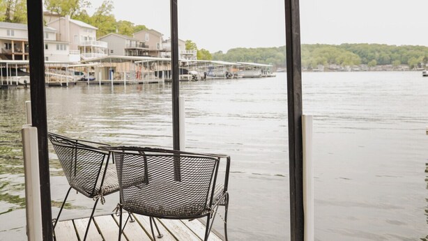 A view of a waterfront property with a covered deck, two metal chairs, and a calm river with boats and buildings in the background. - Waterfront Cottage & Dock w/ Beautiful View - 5MM (Rocky Mount)