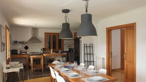 Ein Essbereich mit einem Holztisch, gedeckt für sechs Personen, umgeben von weißen Stühlen. Im Hintergrund ist eine Küche mit modernen Geräten und einem großen Fenster zu sehen.