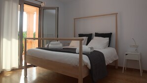 Ein Schlafzimmer mit einem hölzernen Bett, weißer Bettwäsche, einem kleinen Nachttisch und Blick durch eine offene Tür.