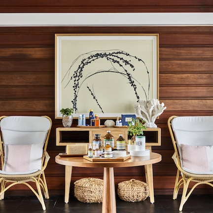 A room with two wicker chairs, a small round table, and a wooden wall with a framed artwork.