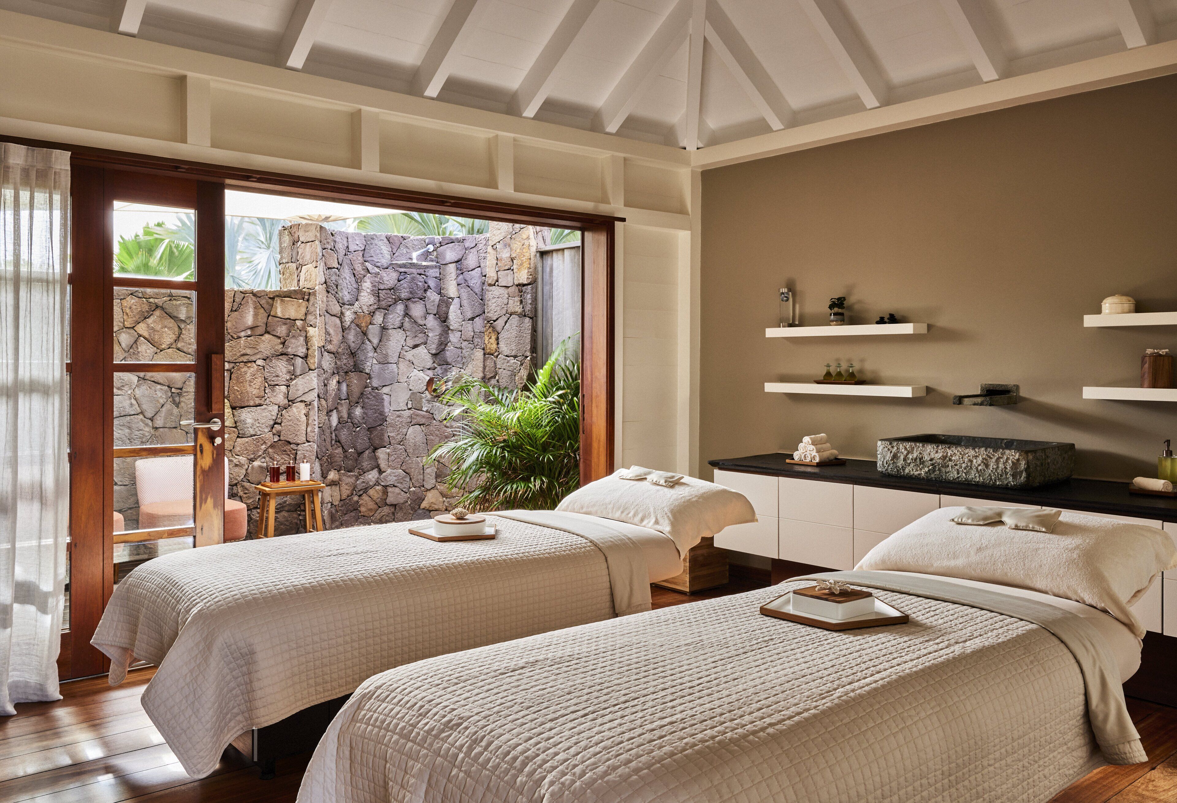 A hotel room with two beds, a stone wall, a small table, and a view of greenery outside.
