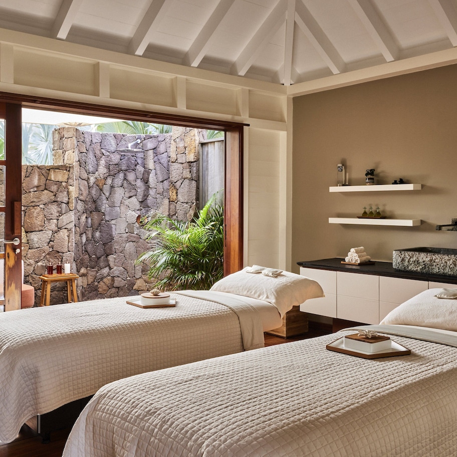 A hotel room with two beds, a stone wall, a small table, and a view of greenery outside.