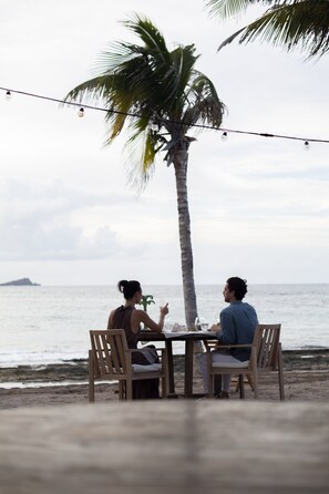Two people seated at a wooden table with a view of the ocean and a palm tree. - Rosewood Le Guanahani St Barth (St. Barthelemy)