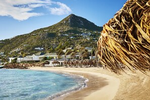 A beach with a mountain in the background, a thatched umbrella, and a building with a white facade. - Rosewood Le Guanahani St Barth (St. Barthelemy)