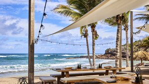 A beachside restaurant with wooden tables and benches, string lights, and palm trees. - Rosewood Le Guanahani St Barth (St. Barthelemy)