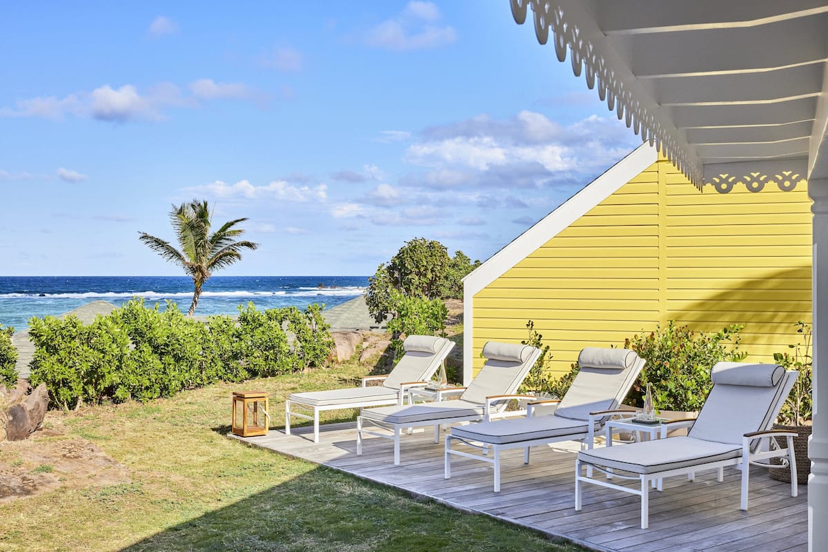 A deck with white lounge chairs and a yellow building with a white roof, overlooking the ocean.