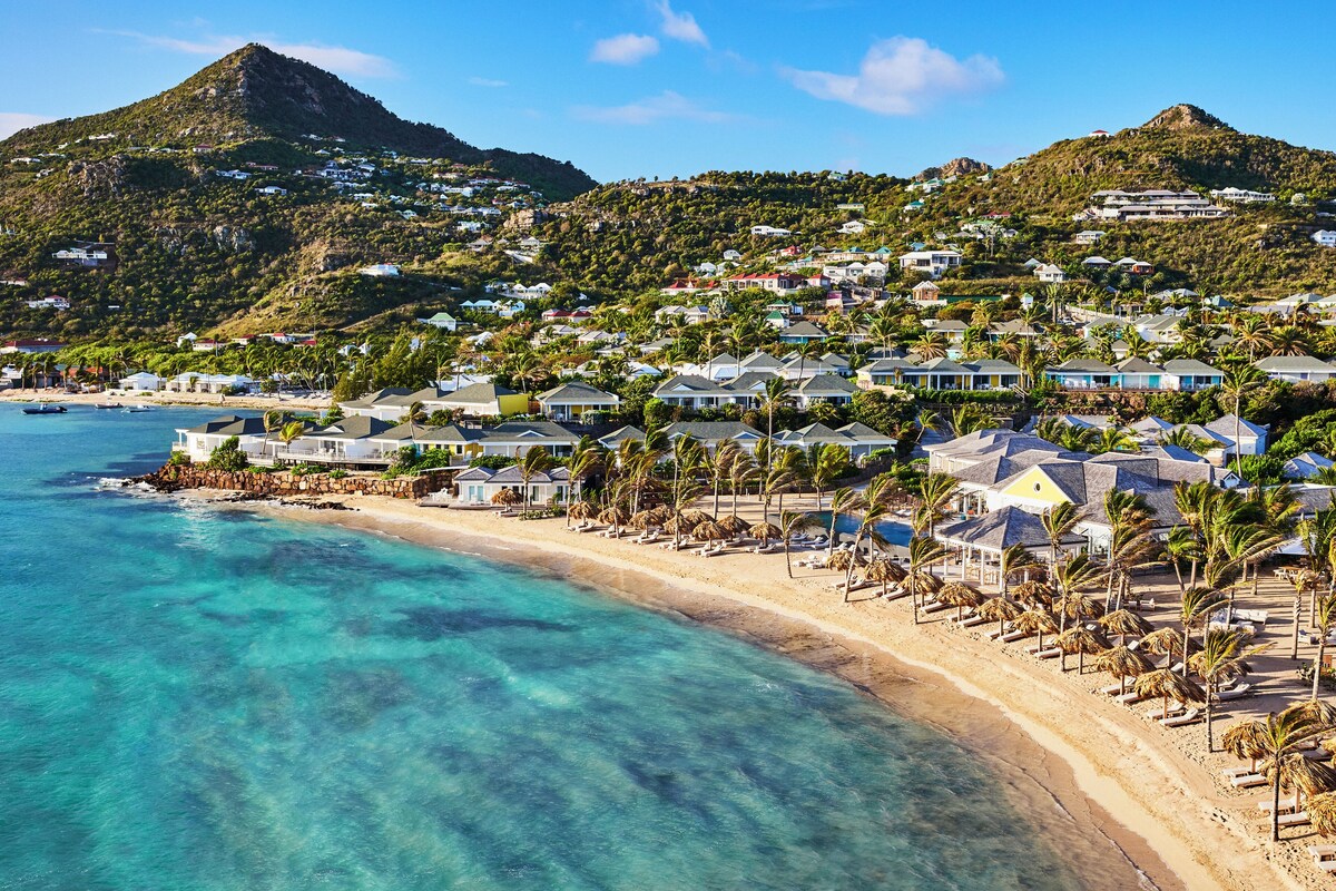 A coastal resort with beachfront villas, clear turquoise waters, and a mountainous backdrop.