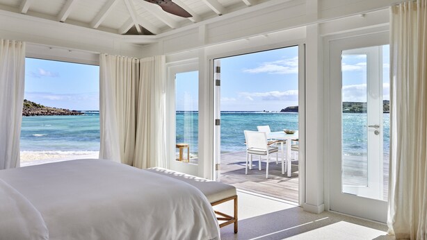 A bedroom with a large bed, a ceiling fan, and a view of the ocean through large windows. - Rosewood Le Guanahani St Barth (St. Barthelemy)