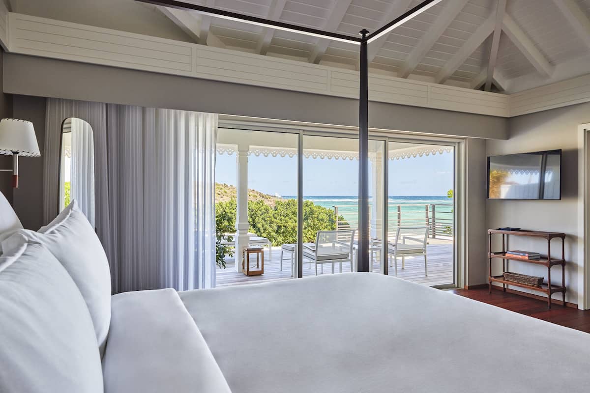 A spacious bedroom with a large bed, a balcony with a view of the ocean, and a television on the wall.