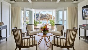 A room with a round table, four chairs, a TV, and a view of the beach and mountains. - Rosewood Le Guanahani St Barth (St. Barthelemy)