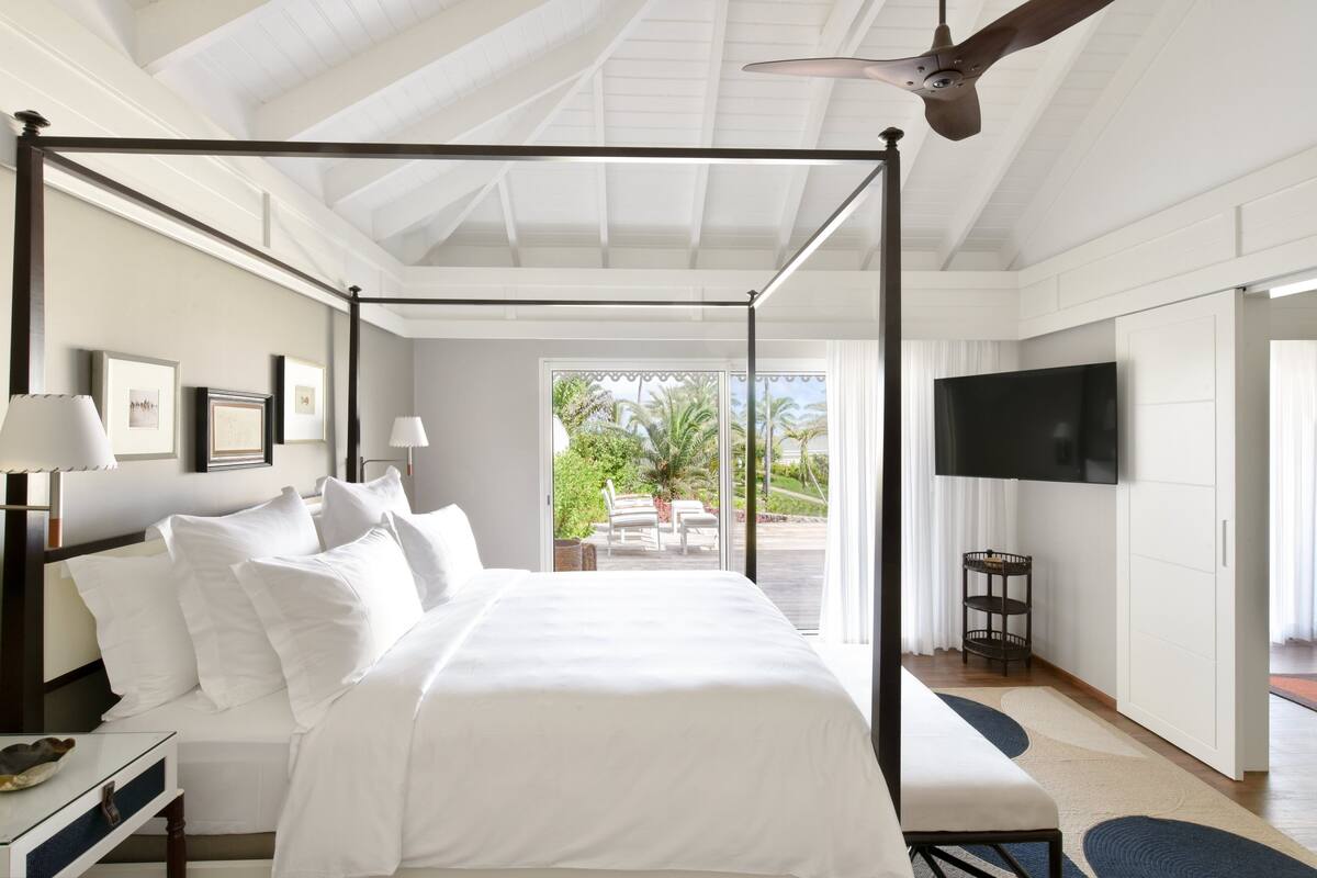 A four-poster bed with white linens, a ceiling fan, a TV on the wall, and a view of the outdoors.