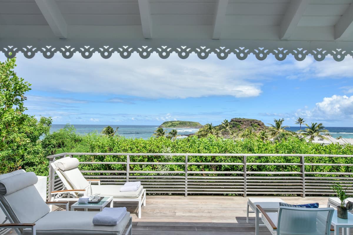 A balcony with white lounge chairs, a wooden deck, and a view of the ocean and a tropical landscape.
