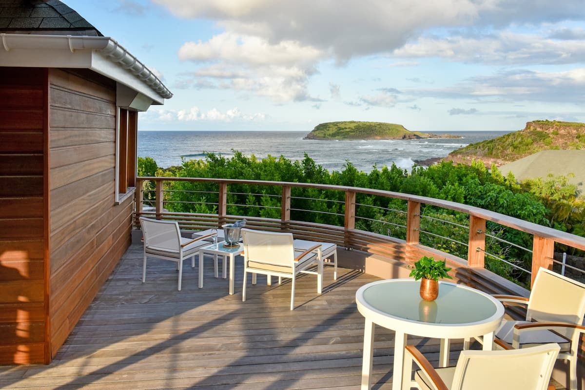 A wooden deck with outdoor furniture, a table with a plant, and a view of the ocean and a distant island.