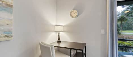 Ein Zimmer mit Schreibtisch, Stuhl, Lampe, Uhr und Blick auf Grünflächen.