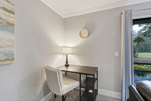 A room with a desk, chair, lamp, clock, and a view of greenery outside.