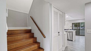 Un escalier avec des marches en bois et une rampe en bois, menant à une pièce dotée de portes blanches et d’un espace cuisine visible à travers une porte en verre.