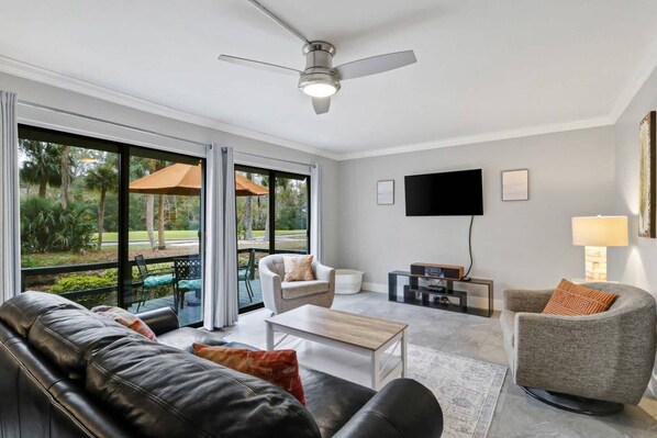 A modern living room with a ceiling fan, a flat-screen TV, and a large window overlooking a garden.