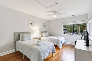 A hotel room with two beds, a desk, a television, and a window with a view of palm trees.