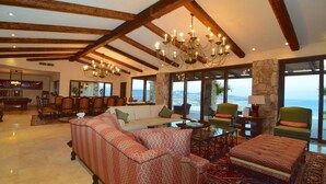 A spacious living room with a large sofa, a coffee table, and a chandelier. There is a dining area with chairs and a table, and large windows offering a view of the ocean. - Luxury Pacific View Villa, Cabo San Lucas Villa 1005 (Cabo San Lucas)