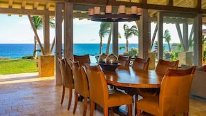 A dining area with a view of the ocean, featuring a wooden table and chairs, a hanging light fixture, and a view of palm trees and the sea. - Your Dream Beach Getaway Awaits, San Jose Del Cabo 1022 (San Jose Del Cabo)