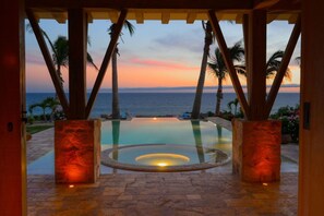 Une piscine à débordement offrant une vue sur l’océan au coucher du soleil, entourée de palmiers et de piliers en pierre.