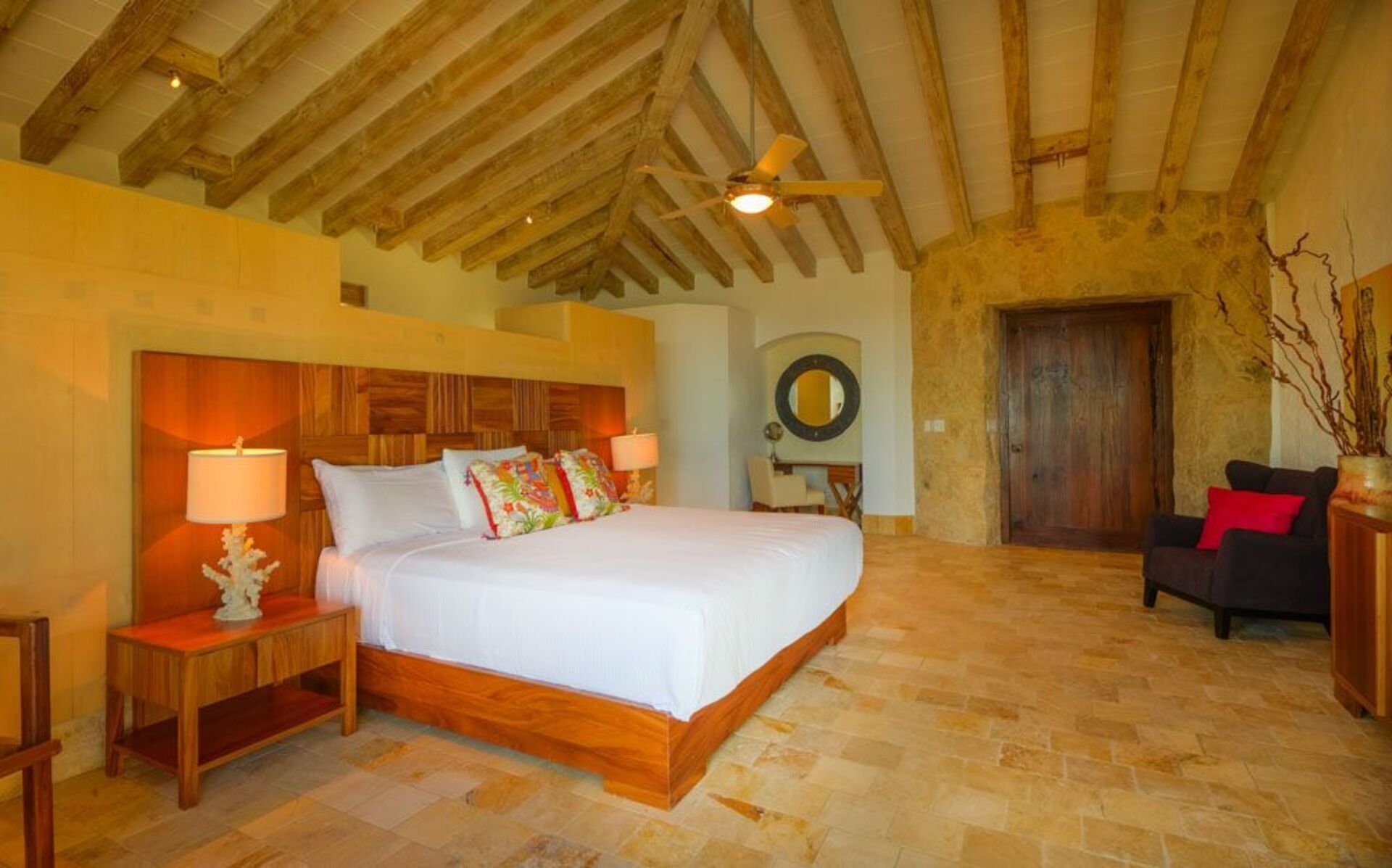 Une chambre à coucher avec un lit en bois, deux tables de chevet avec des lampes, un miroir rond, un canapé foncé et une porte en bois.