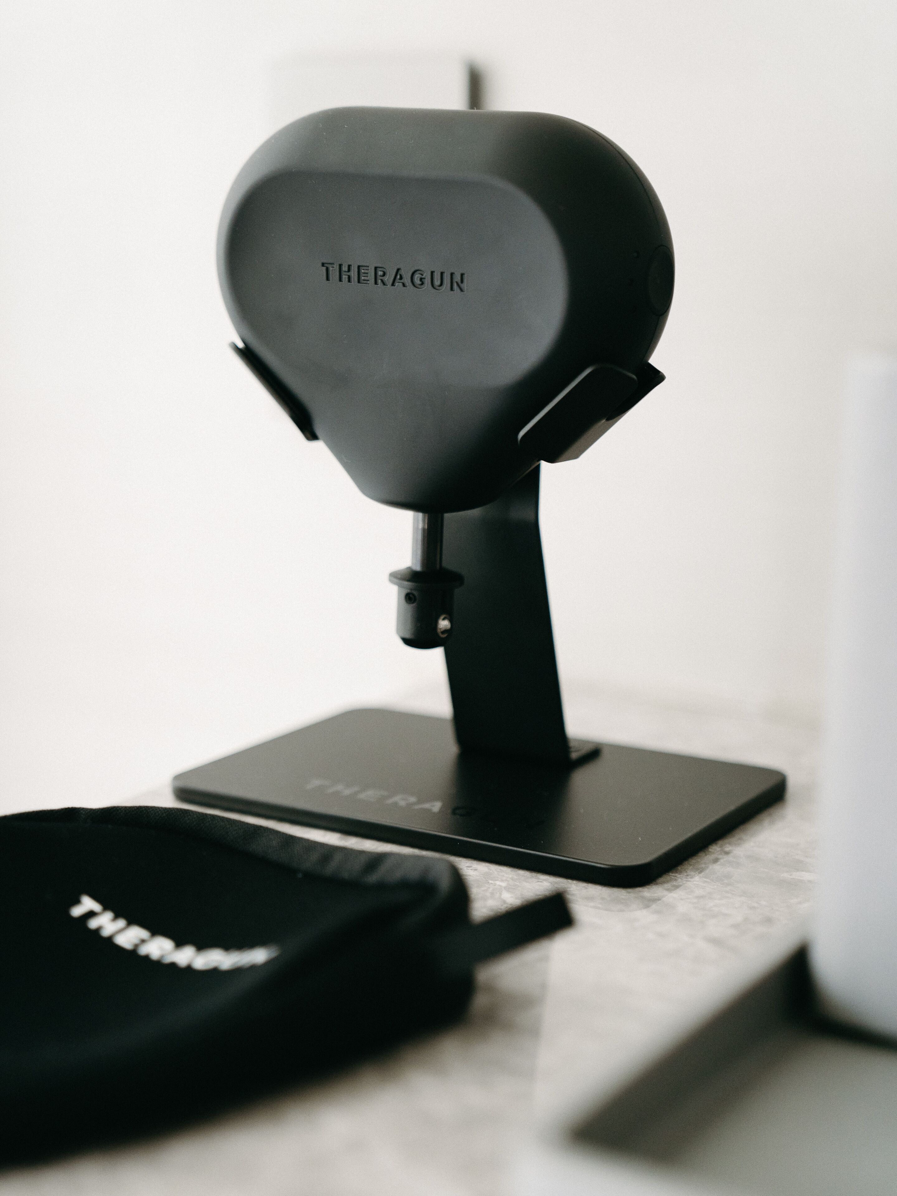 A black THERAGUN device on a stand with a black bag beside it.