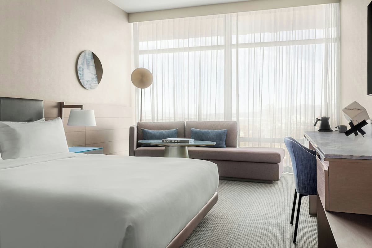 A hotel room with a large bed, a sofa, a desk, and a large window with sheer curtains.