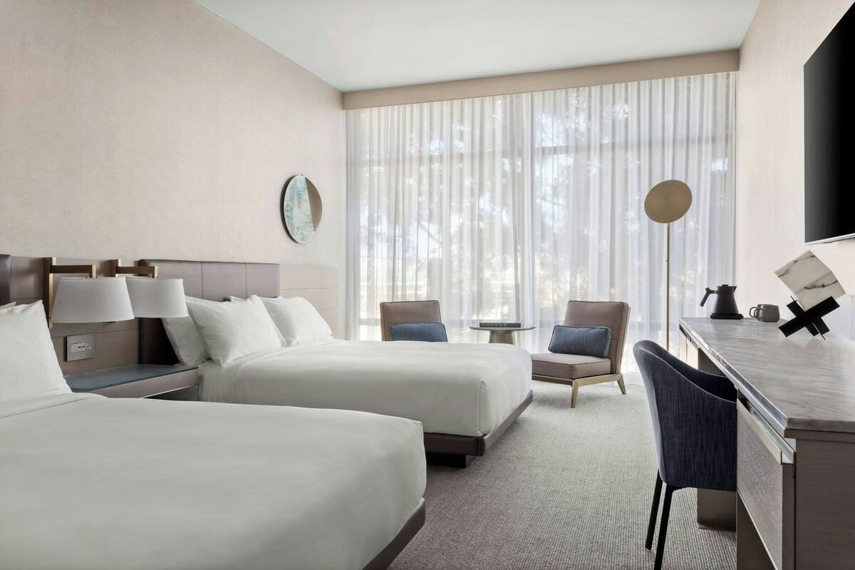A hotel room with two beds, a desk, a chair, a small table, and a large window with curtains.